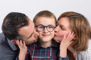 Portrait de famille en studio : des parents embrassent tendrement leur petit garçon à lunettes.