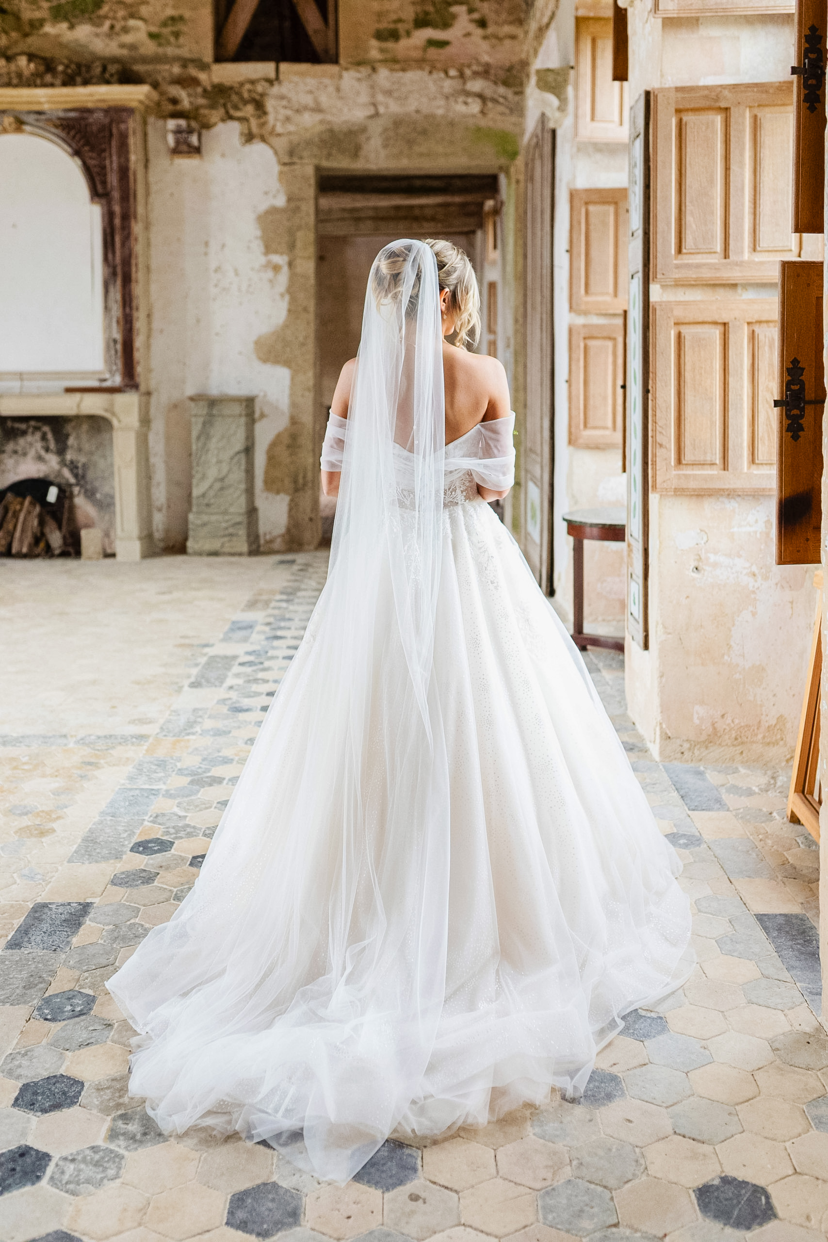 La mariée vue de dos marchant dans un couloir au sol en carreaux anciens, dévoilant la traîne de sa robe et son long voile.