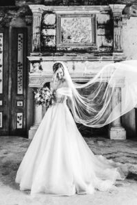 Portrait en noir et blanc de la mariée souriante tenant son bouquet devant une cheminée ancienne monumentale, son voile flottant gracieusement sur le côté.