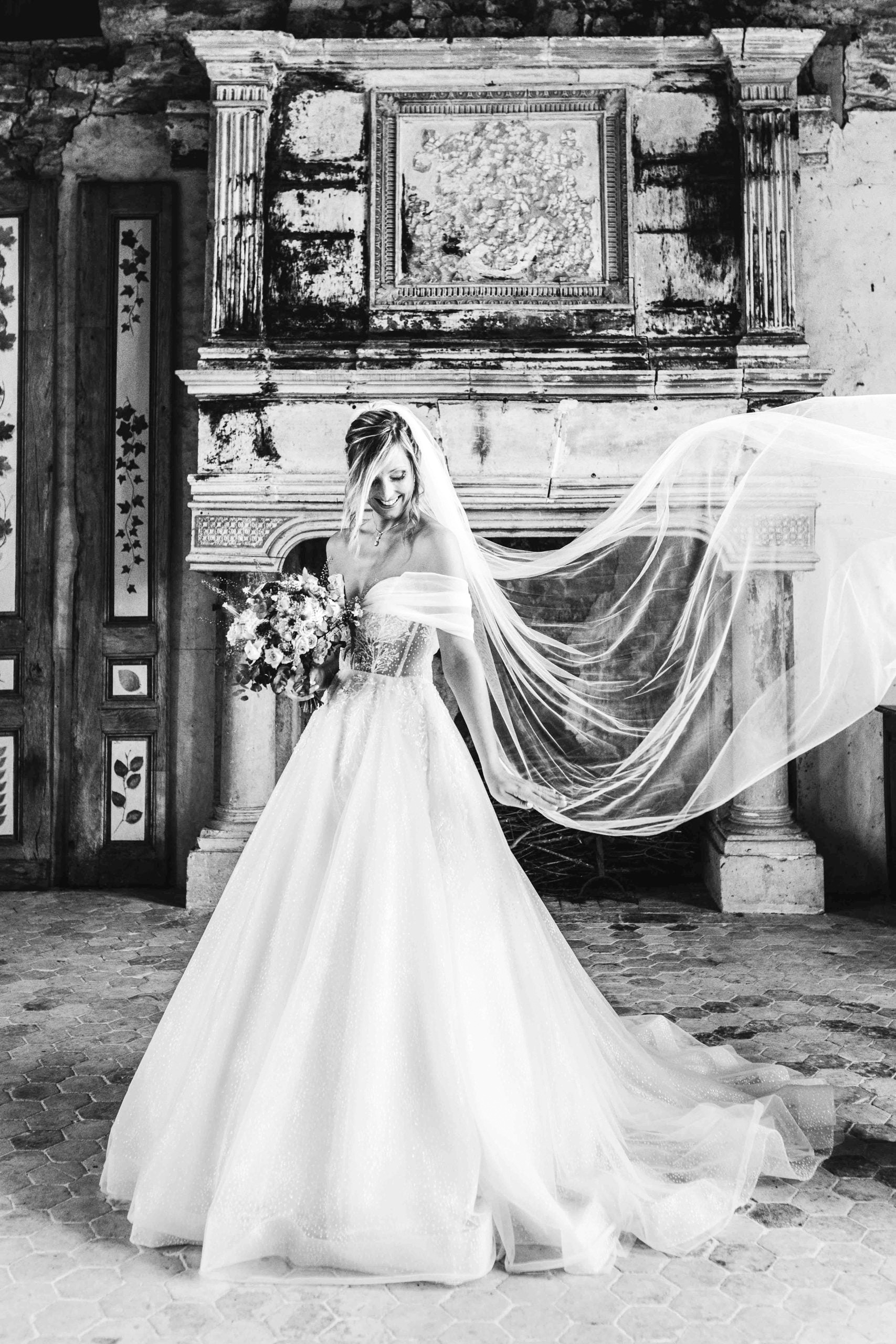 Portrait en noir et blanc de la mariée souriante tenant son bouquet devant une cheminée ancienne monumentale, son voile flottant gracieusement sur le côté.