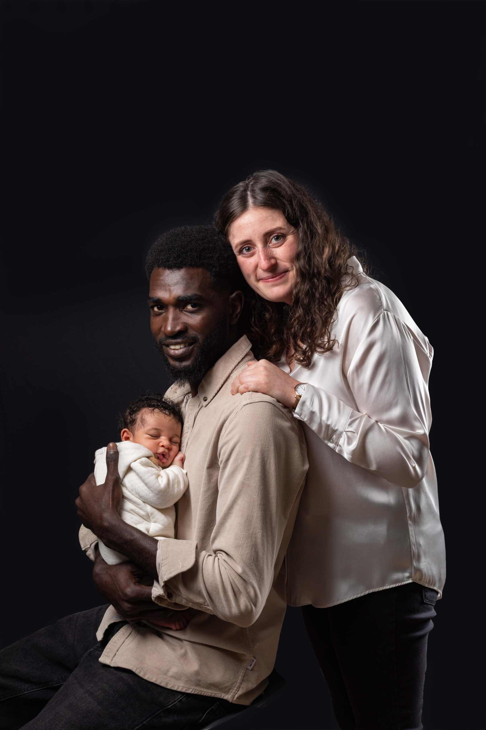 Photographe portraitiste à Deauville : couple posant fièrement avec leur bébé sur fond noir.