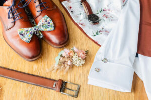 Accessoires du marié disposés sur une table en bois : chaussures en cuir marron, nœud papillon à motifs floraux, montre, boutons de manchette, ceinture et boutonnière en fleurs séchées.