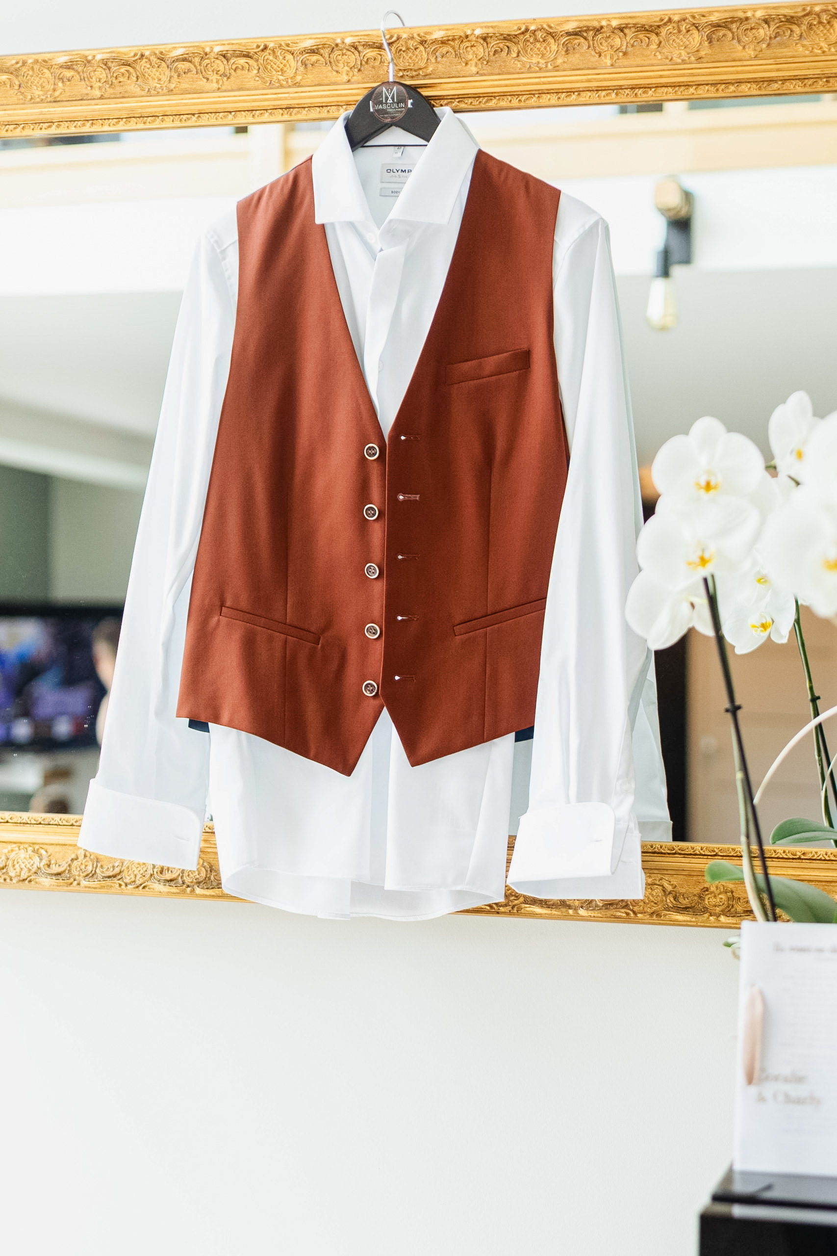 Gilet de costume couleur terracotta et chemise blanche de marié suspendus à un miroir doré, avec une orchidée blanche au premier plan.