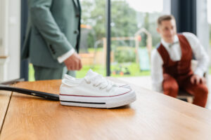 Paire de baskets blanches en toile posée sur une table en bois, avec le marié et un témoin flous en arrière-plan lors des préparatifs.