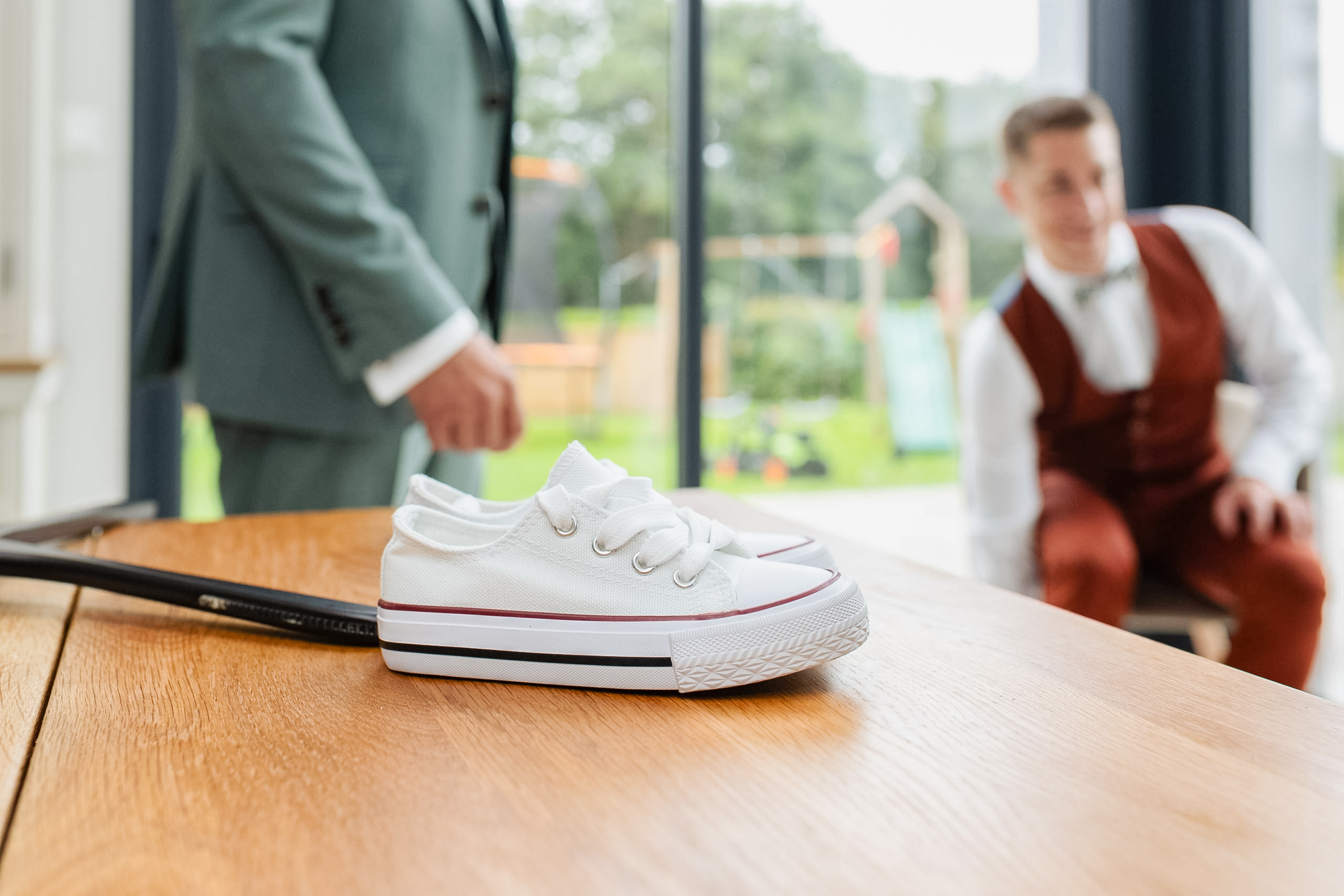 Paire de baskets blanches en toile posée sur une table en bois, avec le marié et un témoin flous en arrière-plan lors des préparatifs.
