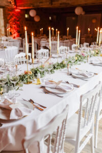 Table de réception de mariage élégante avec nappe blanche, chemin de table végétal en eucalyptus, grands chandeliers dorés, vaisselle délicate et chaises blanches.