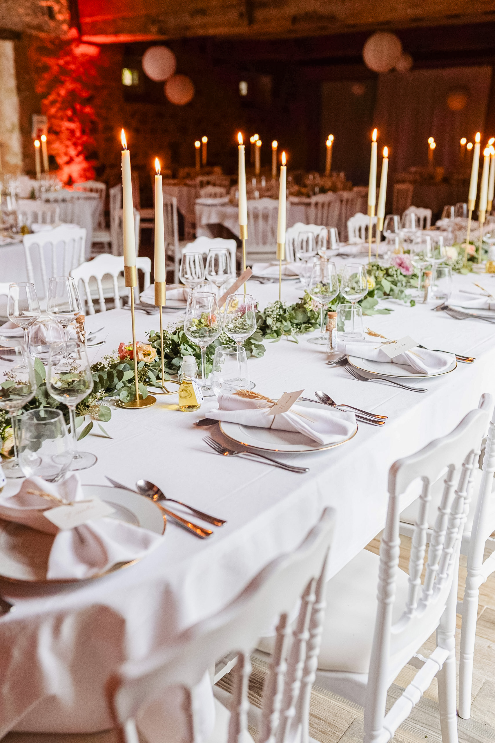 Table de réception de mariage élégante avec nappe blanche, chemin de table végétal en eucalyptus, grands chandeliers dorés, vaisselle délicate et chaises blanches.
