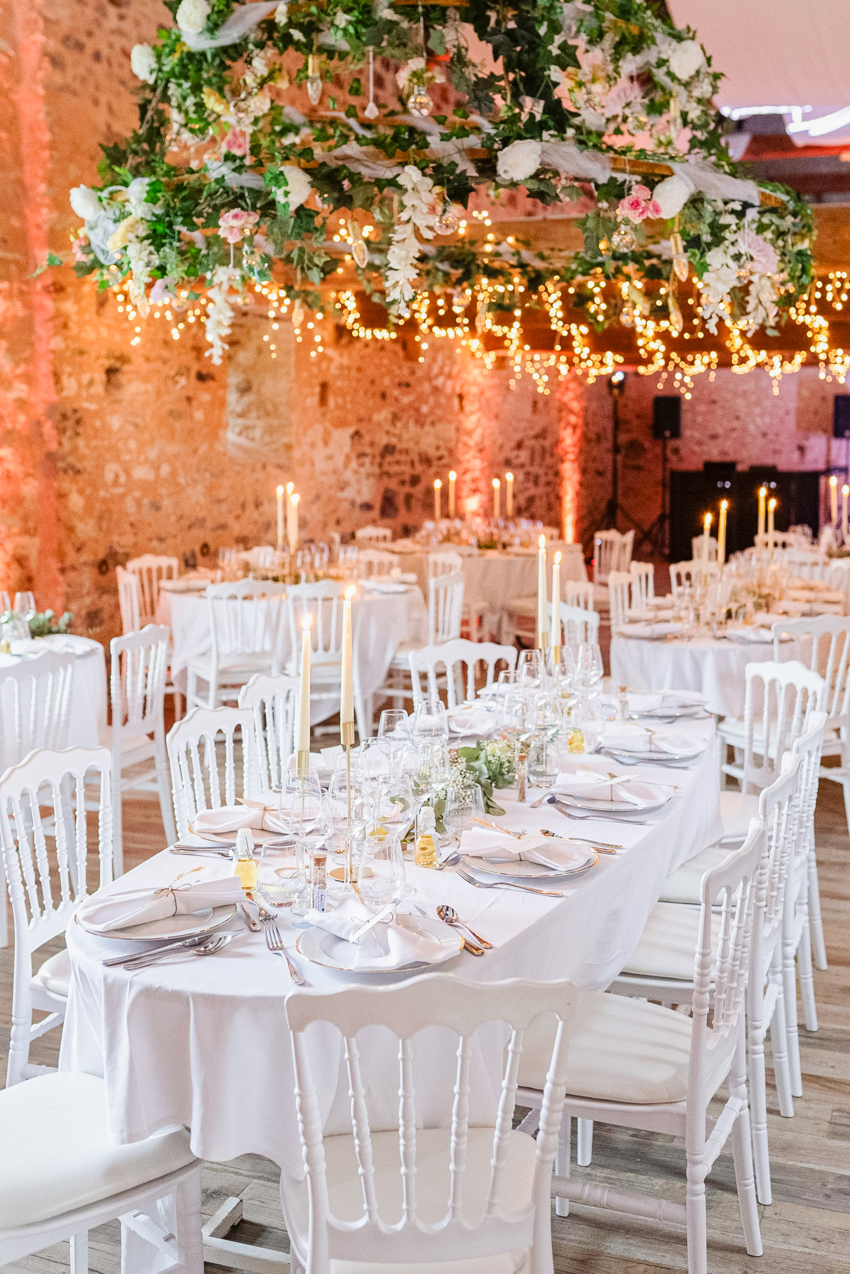 Décoration d'une salle de mariage dans une grange en pierres, avec des tables rondes, un éclairage chaleureux et un grand lustre végétal et floral suspendu au plafond.