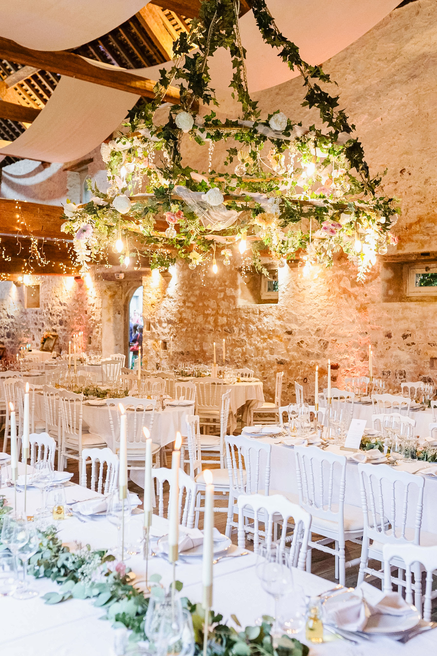 Longue table de banquet de mariage dressée avec de la vaisselle blanche, des bougies fines et une décoration florale, sous un plafond en pierres apparentes avec un lustre en lierre.