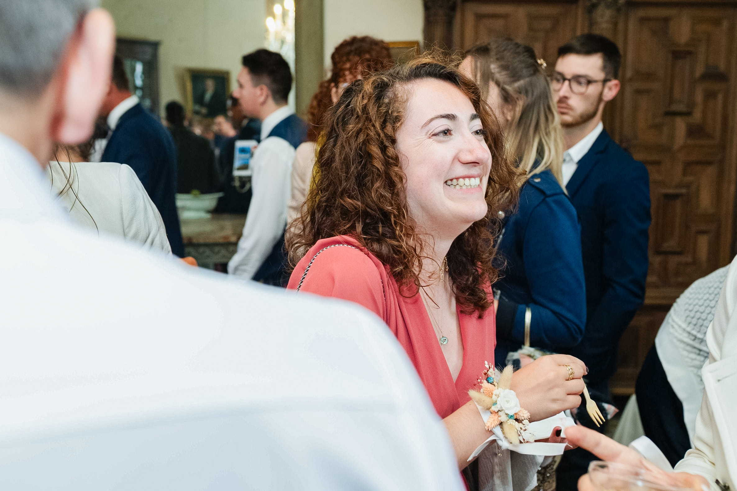 Portrait d'une invitée de mariage souriante portant une robe corail et un bracelet fleuri au poignet lors du vin d'honneur.
