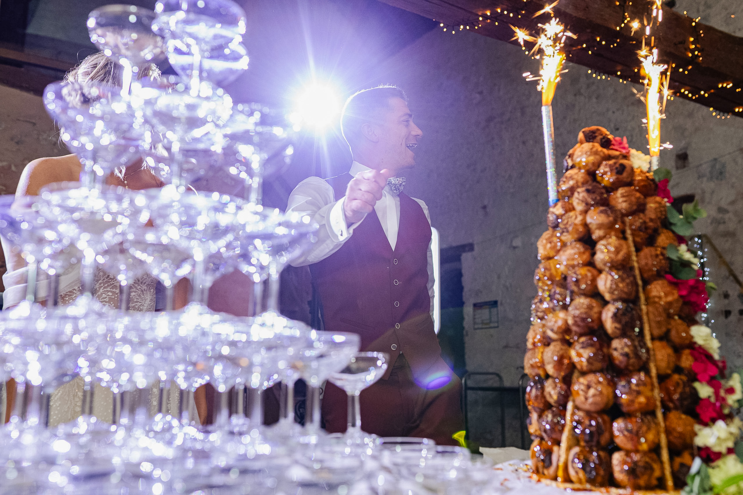 Les mariés lèvent les bras en signe de joie derrière leur pièce montée et leur fontaine à champagne lors de la soirée, avec le DJ en arrière-plan.
