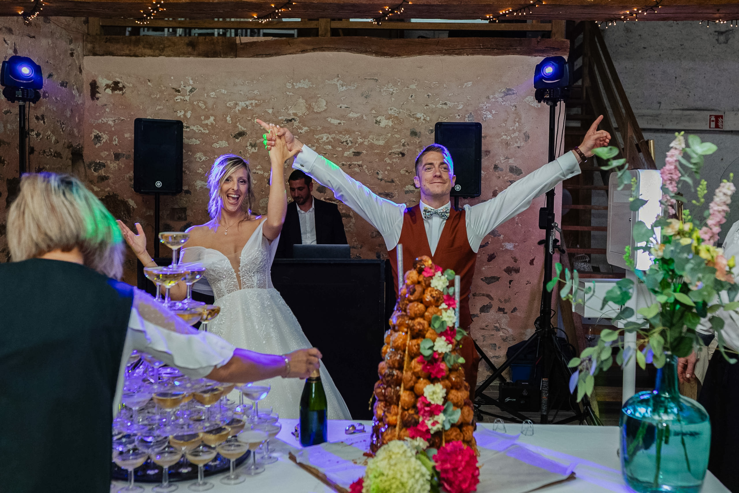 Le marié souriant et pointant du doigt derrière une pièce montée en choux surmontée de cierges magiques, avec une pyramide de verres à champagne floue au premier plan.