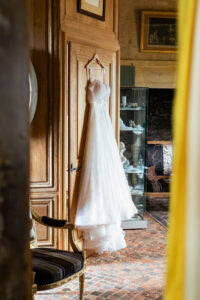 Robe de mariée blanche bustier à paillettes suspendue à un cintre sur une porte en bois sculpté, dans une pièce au décor antique.