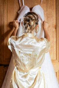 La mariée vue de dos portant un peignoir en satin doré avec l'inscription brillante "Coralie LA MARIÉE", ajustant sa robe de mariée suspendue devant elle.