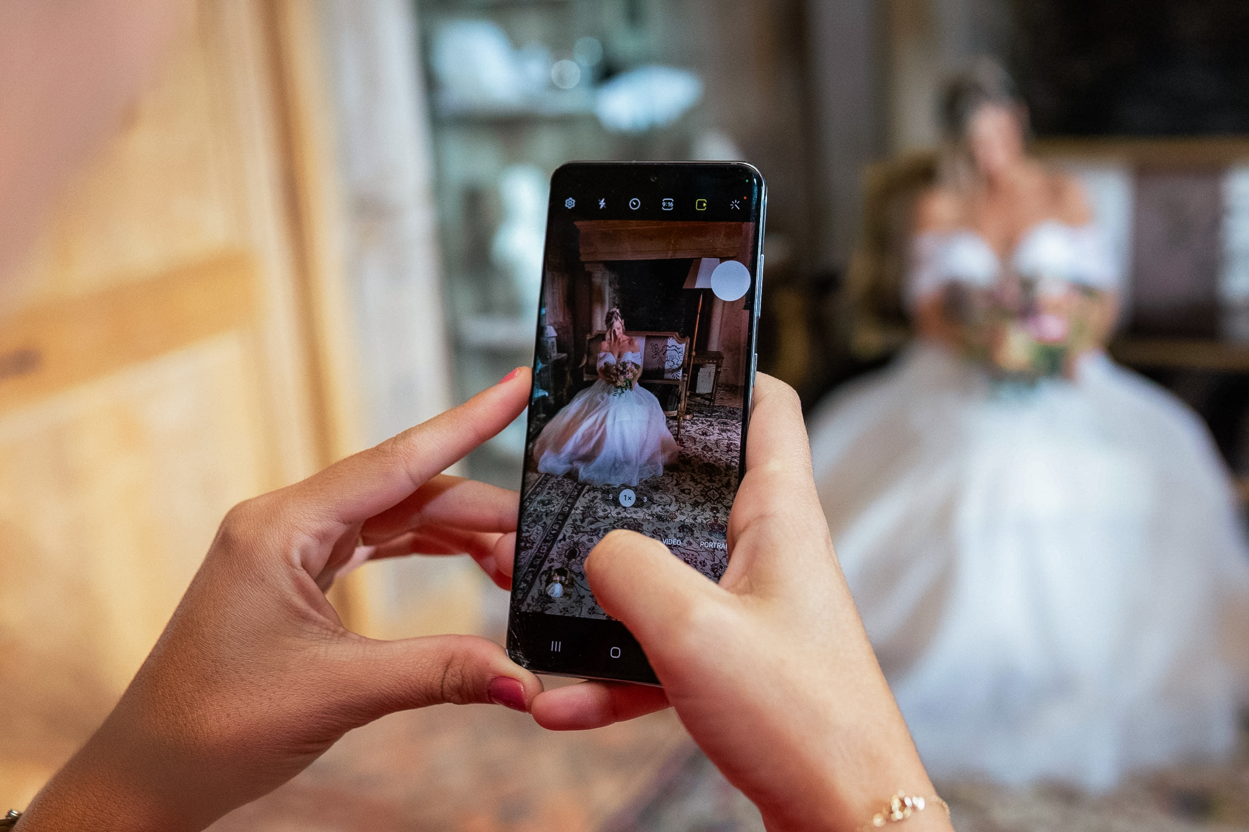 Gros plan sur un écran de smartphone prenant en photo la mariée assise avec son bouquet ; la véritable mariée apparaît floue en arrière-plan.