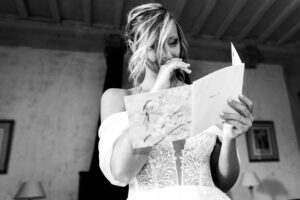 Portrait en noir et blanc de la mariée très émue, la main sur la bouche, lisant une carte de vœux lors de ses préparatifs.