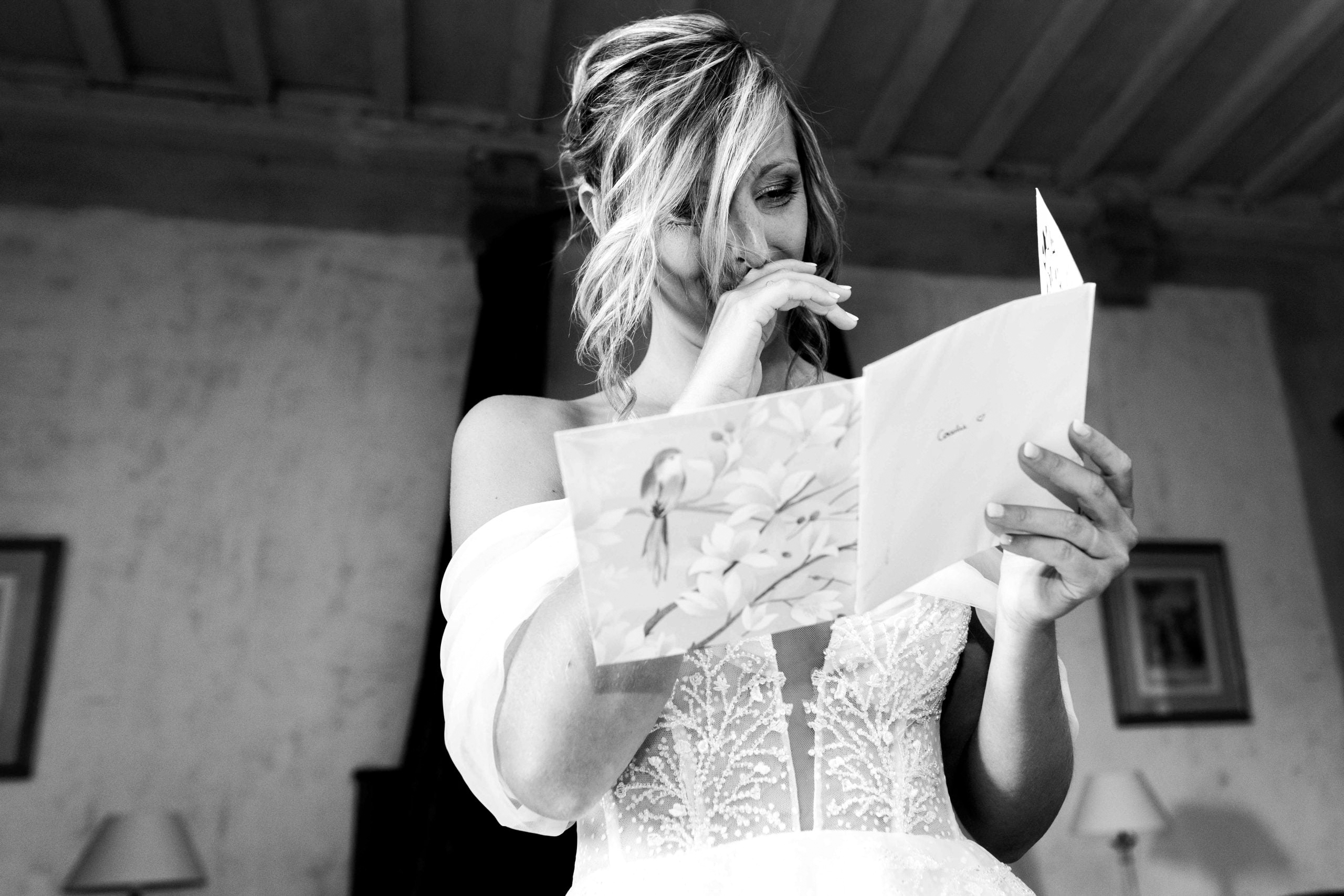 Portrait en noir et blanc de la mariée très émue, la main sur la bouche, lisant une carte de vœux lors de ses préparatifs.