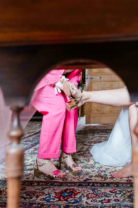 Vue originale sous un meuble ancien montrant une personne en tailleur rose fuchsia aidant la mariée à enfiler ses sandales à talons dorées sur un tapis persan.
