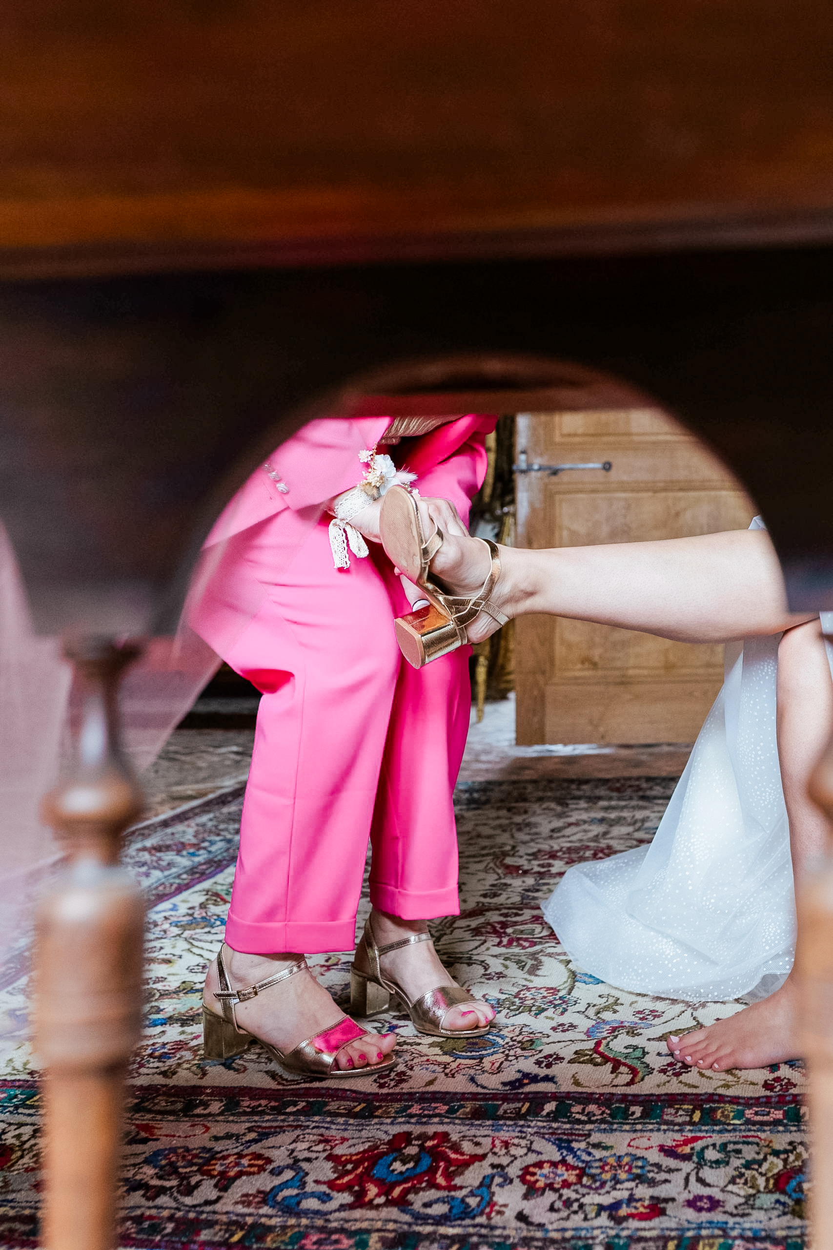 Vue originale sous un meuble ancien montrant une personne en tailleur rose fuchsia aidant la mariée à enfiler ses sandales à talons dorées sur un tapis persan.