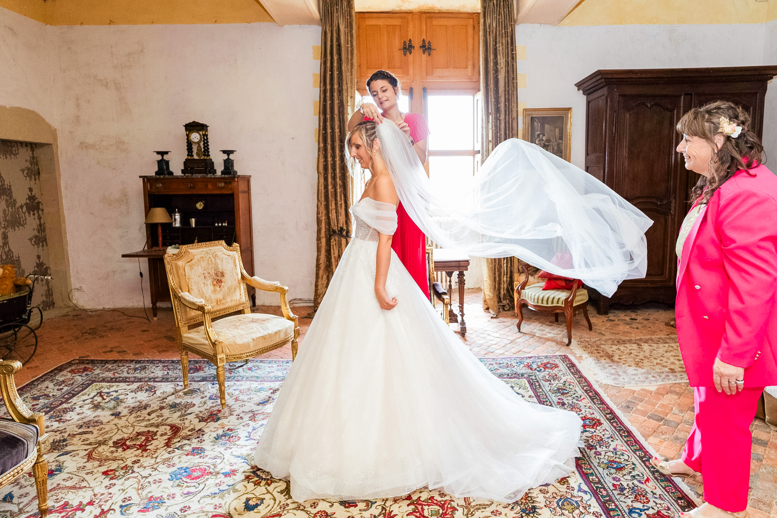 Une amie en robe rouge ajuste le voile de la mariée en le faisant voler au centre d'une pièce ancienne, sous le regard souriant d'une femme en veste rose.