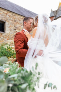 : Portrait romantique des mariés s'enlaçant devant un bâtiment en pierre, le long voile en tulle de la mariée volant au vent au premier plan.