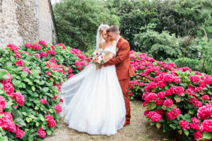 Le marié embrasse tendrement la mariée sur la joue au milieu d'une allée de jardin bordée de magnifiques hortensias roses éclatants.