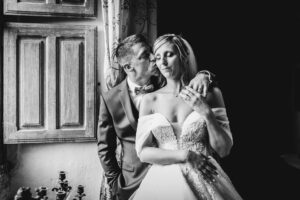 Tendre portrait en noir et blanc du marié déposant un baiser sur la joue de la mariée, les yeux fermés et apaisée, près d'une fenêtre à volets en bois.