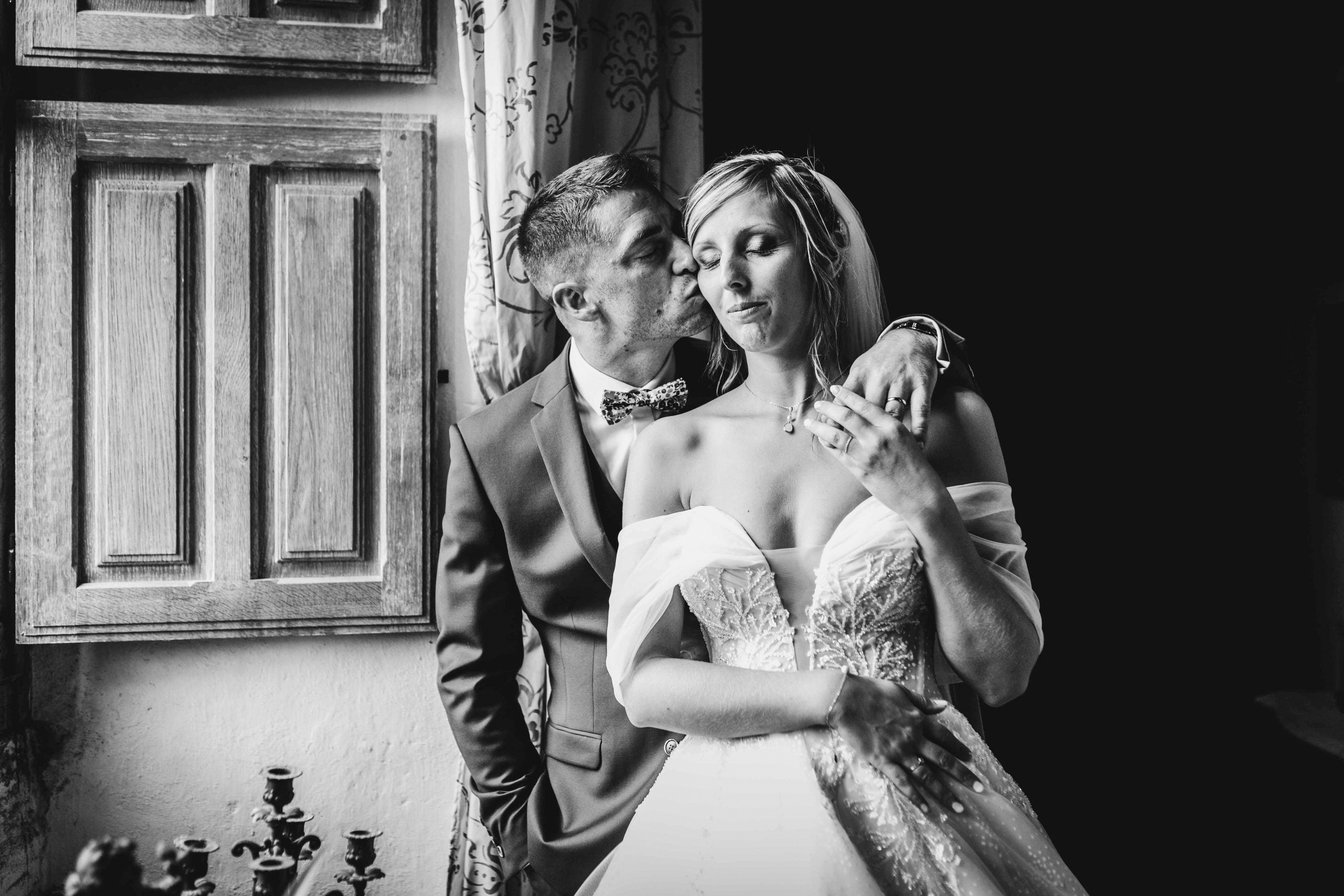 Tendre portrait en noir et blanc du marié déposant un baiser sur la joue de la mariée, les yeux fermés et apaisée, près d'une fenêtre à volets en bois.