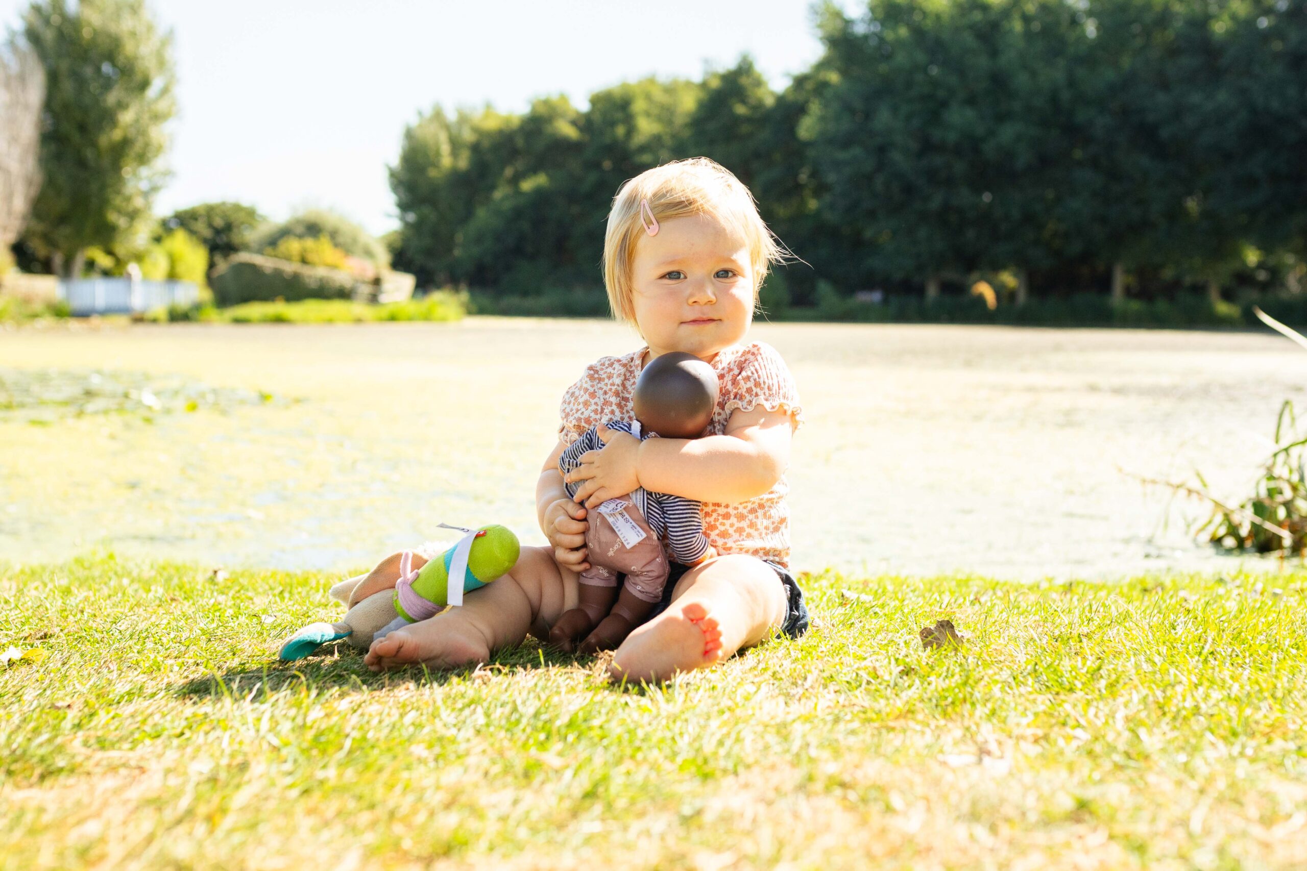 Une petite fille blonde assise dans l'herbe au bord d'un lac, serrant tendrement sa poupée contre elle.