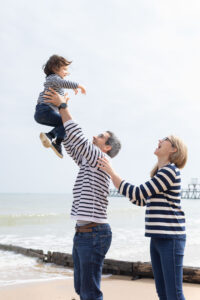 Un père lançant son enfant en l'air sur la plage, accompagné d'une maman souriante, tous habillés en marinières.