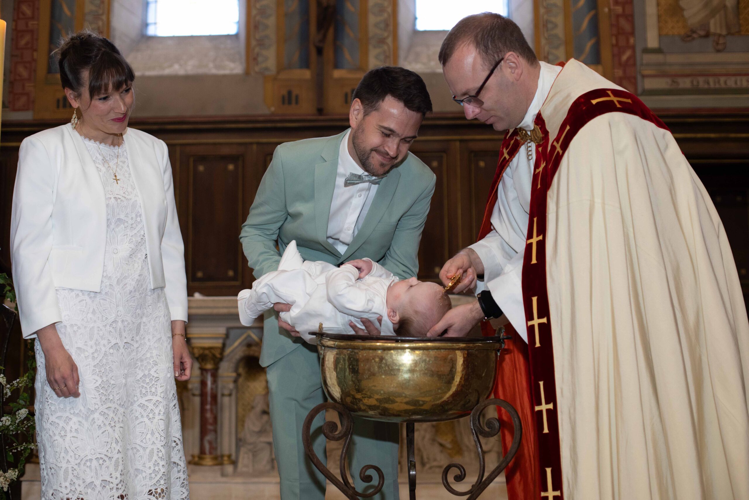Un prêtre verse de l'eau bénite sur la tête d'un bébé lors d'un baptême à l'église, entouré de ses parents émus.