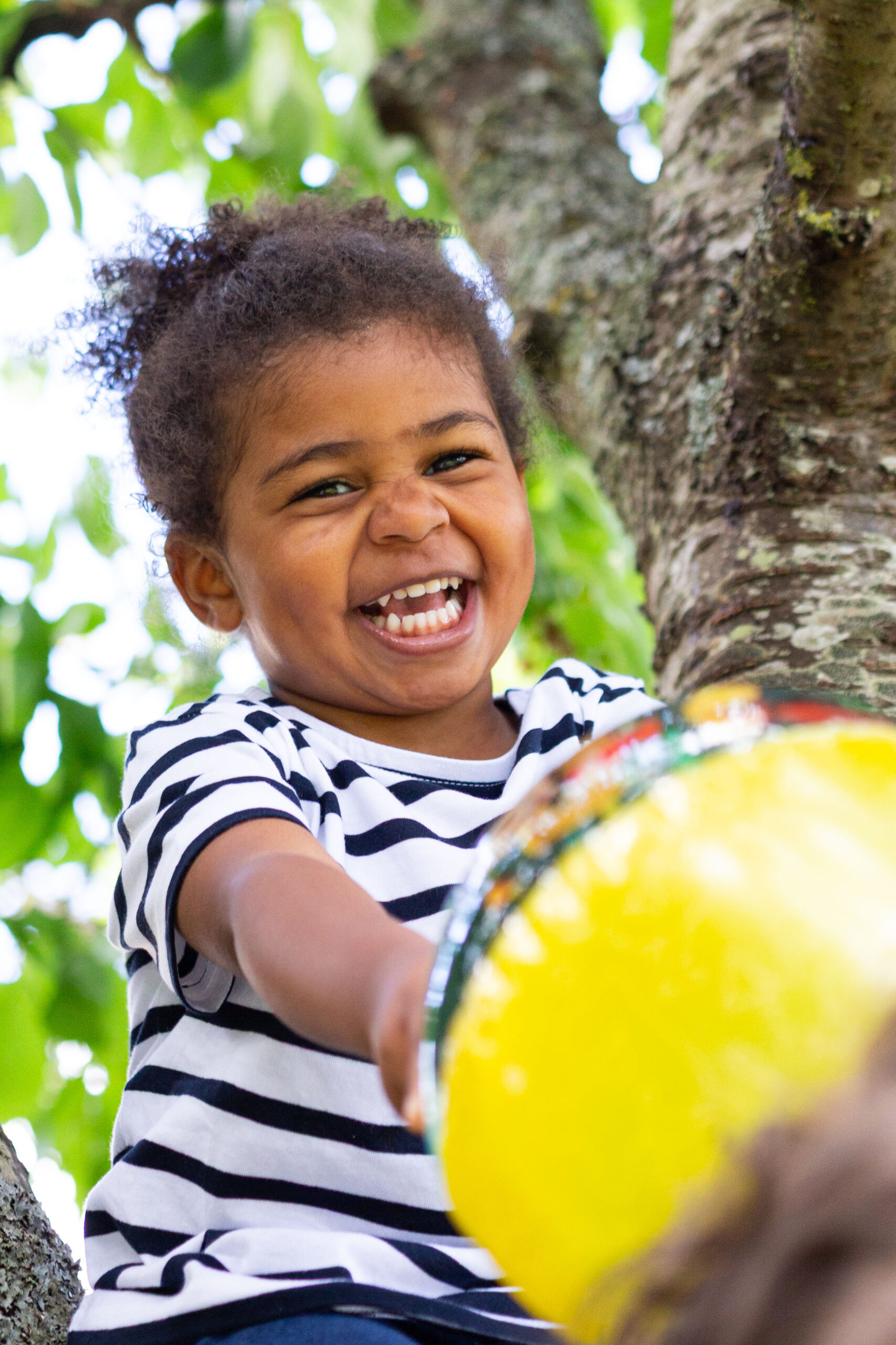 Portrait rapproché d'une petite fille métisse aux cheveux bouclés, affichant un grand sourire radieux en extérieur.