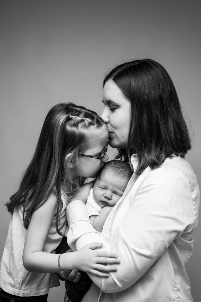 Photographie en noir et blanc d'une maman et de la grande sœur embrassant tendrement le nouveau-né, photographe naissance Le Havre.