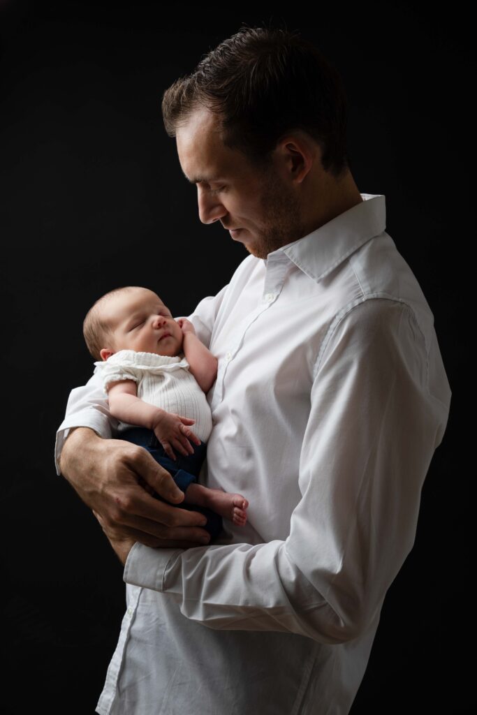 Portrait de naissance en studio sur fond noir : un jeune papa berce son bébé endormi, photographe nouveau-né Deauville.