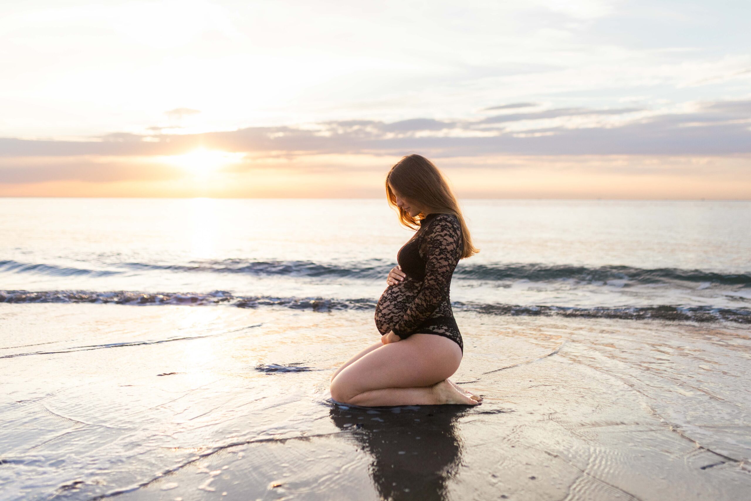 Femme enceinte posant à genoux sur le sable au coucher du soleil en body dentelle noir, photographe grossesse près de Deauville.